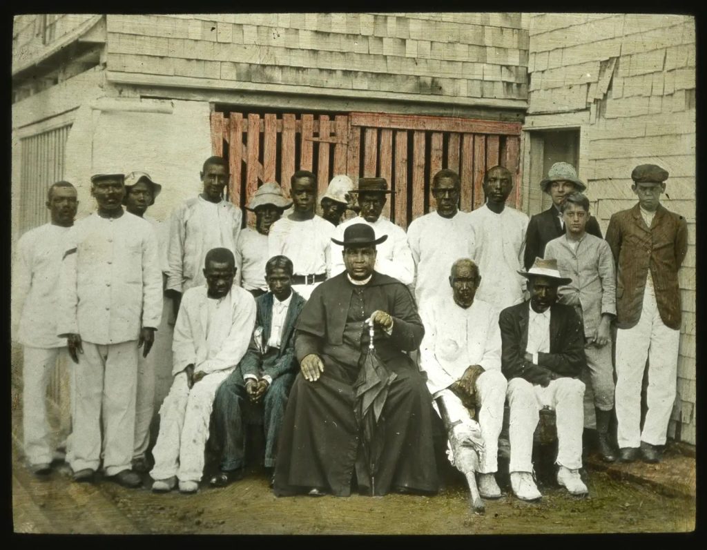 A formally arranged group outside a timber-clad institutional or communal building in Jamaica. The central religious figure, uniformity of dress and enclosed façade suggest an organised mission, hospital, workhouse or similar institution, although the precise setting and identities require archival confirmation. The image offers a reminder that colonial buildings also organised authority, welfare, religion and social control.