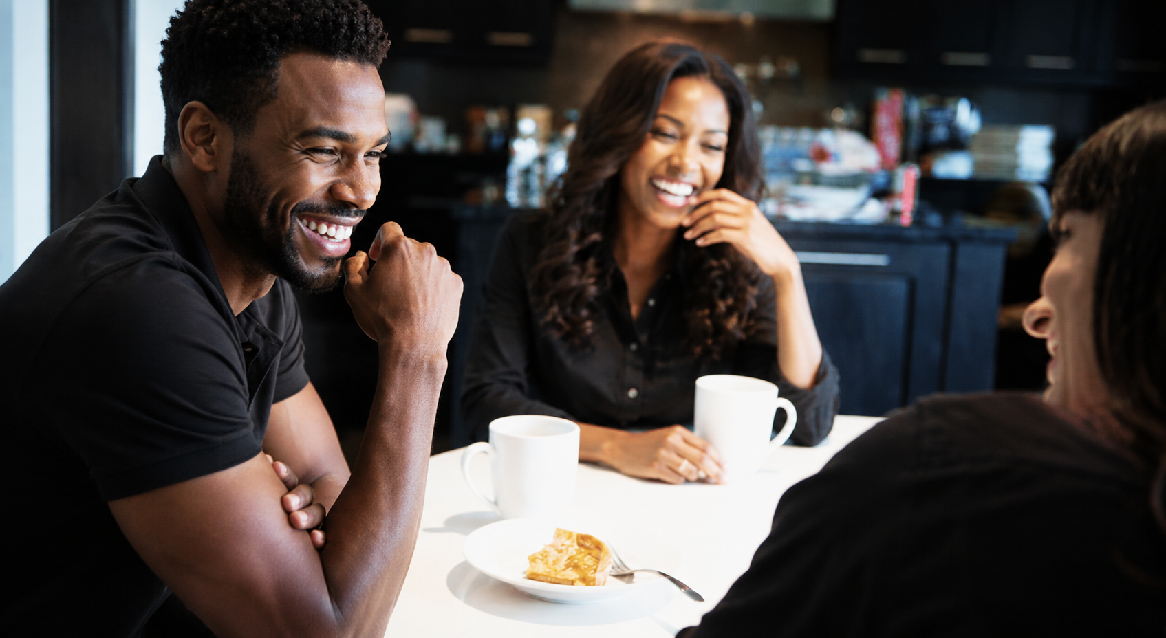A warm, candid kitchen scene showing two Black couples laughing together around a table with coffee mugs and dessert. The group mirrors the relaxed poses and joyful expressions of a casual lifestyle photo, with soft lighting, dark modern cabinetry, and a cozy, friendly atmosphere.