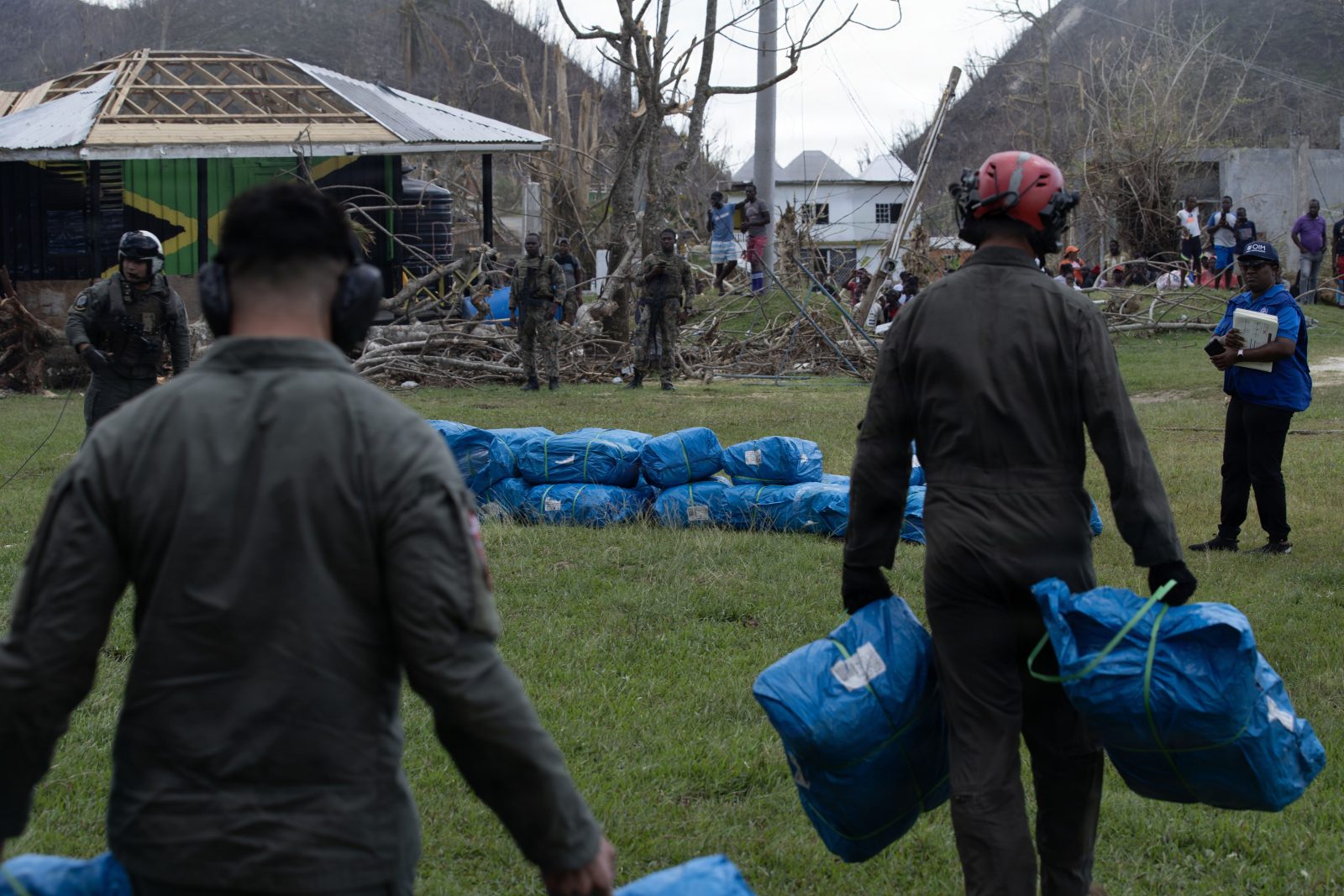 Shelter supplies in blue bags being unloaded in Beeston Spring, Jamaica, beside a house that lost its roof in Hurricane Melissa