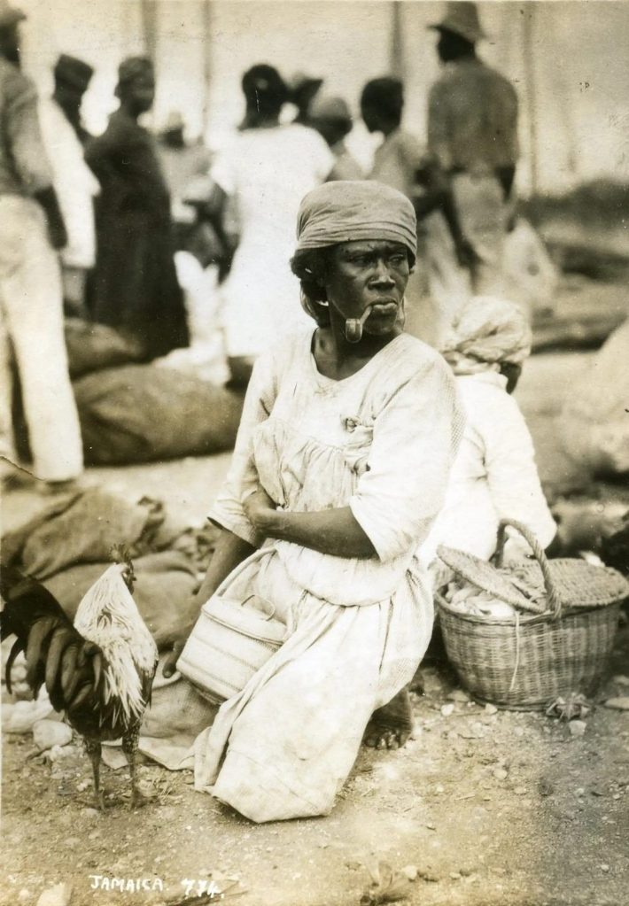 A Jamaican market woman photographed with baskets, bags and poultry, probably during the late 19th or early 20th century. Her portable stock illustrates how women’s labour connected small farms, roadside commerce and the growing towns. The image’s exact date should be verified before publication.
