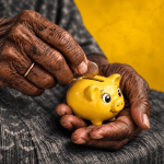 An elderly Jamaican woman’s deeply lined, weathered hands carefully place a coin into a bright yellow piggy bank, capturing the quiet financial pressure facing people who may enter retirement still paying for housing. The dark grey surroundings and aged hands contrast sharply with the cheerful yellow piggy bank, symbolising the difficult balance between limited retirement income, rising living costs and the need to keep saving for security later in life.