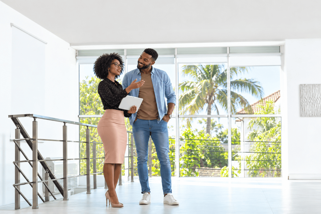 A professional Black Jamaican real estate agent with natural afro hair, glasses and a fitted pink pencil skirt shows a modern property to a male client. Holding a tablet and gesturing towards the home's features, she remains focused on the viewing, while the casually dressed man smiles admiringly at her. Large windows, contemporary interiors and tropical palm trees provide the backdrop for a scene exploring the blurred lines between professional courtesy, personal attraction and inappropriate attention in the property industry.