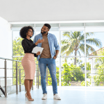 A professional Black Jamaican real estate agent with natural afro hair, glasses and a fitted pink pencil skirt shows a modern property to a male client. Holding a tablet and gesturing towards the home's features, she remains focused on the viewing, while the casually dressed man smiles admiringly at her. Large windows, contemporary interiors and tropical palm trees provide the backdrop for a scene exploring the blurred lines between professional courtesy, personal attraction and inappropriate attention in the property industry.