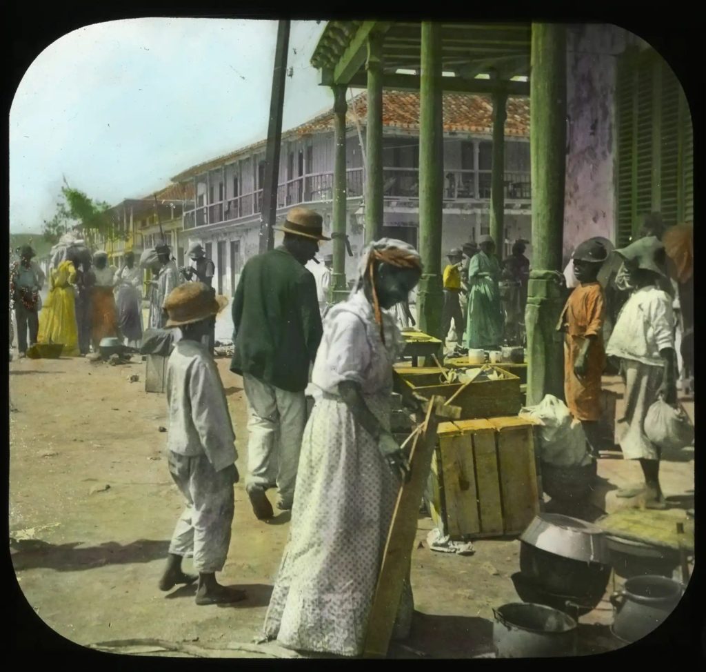 Street trading beneath arcaded and balconied buildings in Jamaica around the turn of the 20th century. The covered walkways offered shade and commercial frontage, while the street itself operated as market, meeting place and transport route. The colour appears to have been applied to the original lantern slide.