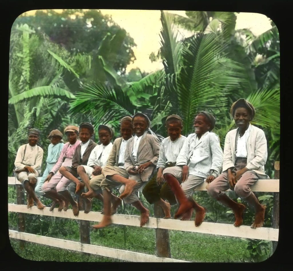 A group of young Jamaican workers pause on a timber fence amid dense tropical planting. Their clothes and bare feet suggest agricultural or estate labour, while their expressions preserve a rare moment of ease within a demanding rural economy. Exact location, occupation and date remain unconfirmed.
