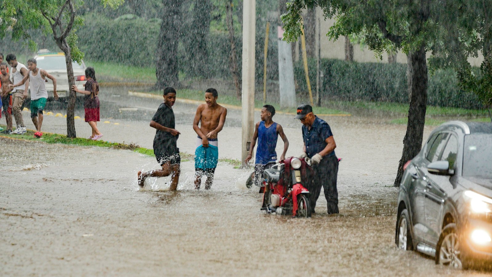 Young men wade through a flooded street in heavy rain while a man pushes a red motorcycle and a car drives past