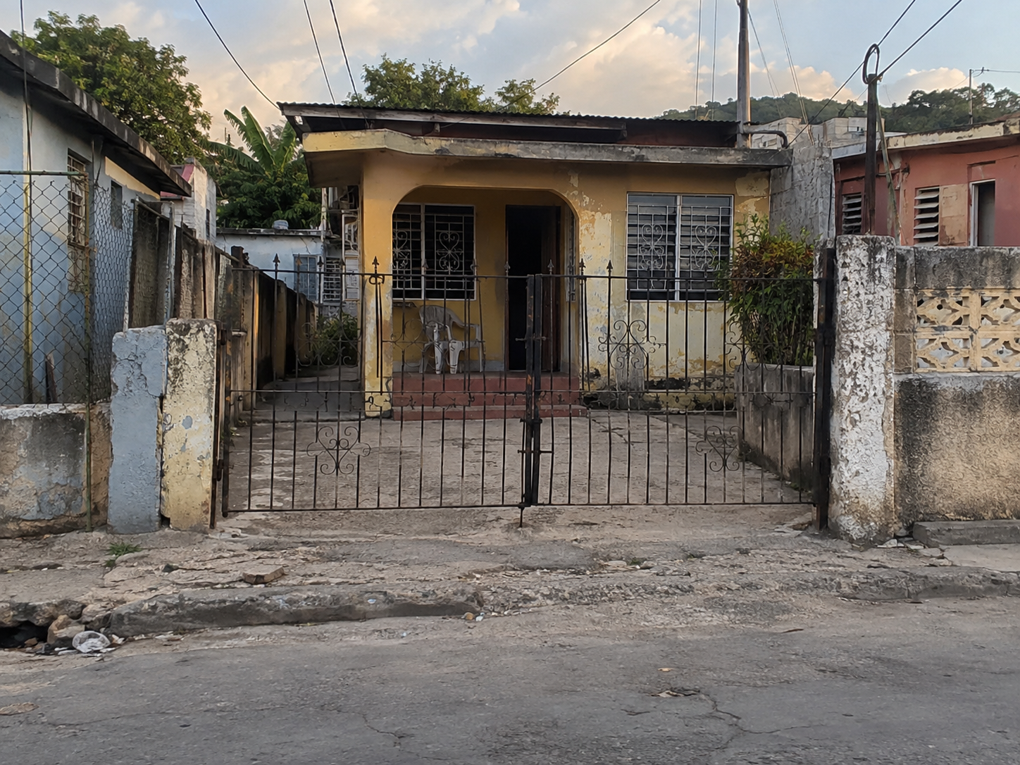 A modest family home behind a secure metal gate in an inner city community in Spanish Town, St Catherine, reflecting the everyday reality of housing, security and neighbourhood life in some of Jamaica’s older urban communities. The weathered exterior, barred windows, narrow residential street and closely built neighbouring homes illustrate how the home remains both a place of shelter and a deeply personal space where families seek safety, stability and belonging.