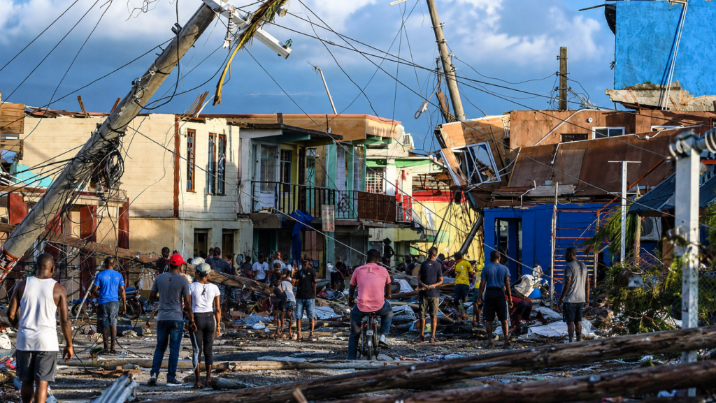 Land Titling Delays Hurricane Housing Scene of widespread destruction following Hurricane Melissa in Jamaica, showing collapsed utility poles, damaged buildings, and residents navigating the debris during early recovery efforts.